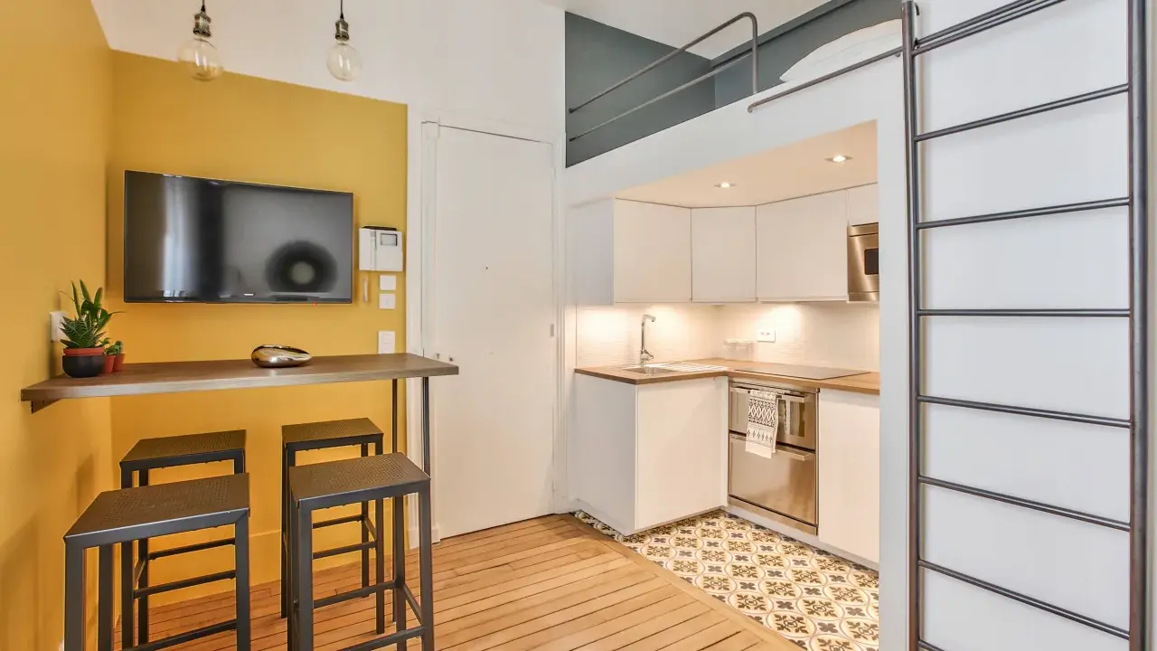 Aménagement studio 18m² : cuisine moderne, coin repas jaune vif avec TV, et lit mezzanine accessible par une échelle.