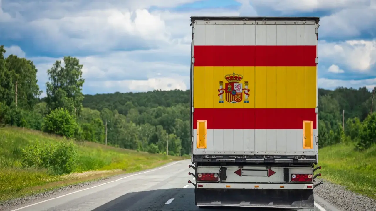 Camion aux couleurs de l'Espagne sur une route de campagne, prêt à déménager en Espagne. Paysage verdoyant sous un ciel nuageux.