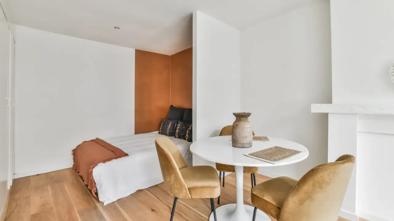 Idée pour aménager studio 30m2 en 2 pièces : un coin nuit cosy avec lit et tête de lit ocre, séparé par une table ronde et des fauteuils velours.