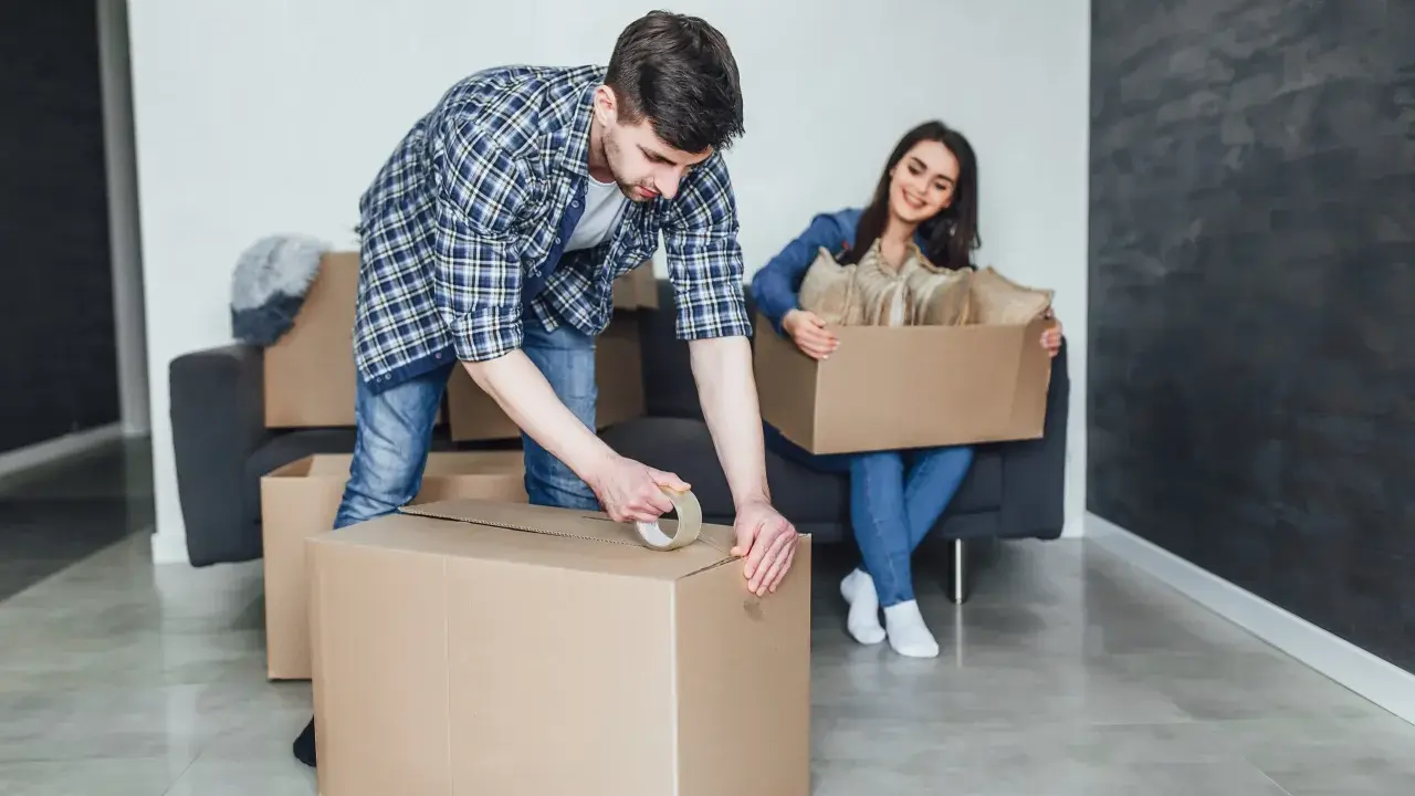 Un homme apprend comment scotcher un carton pour déménager, tandis qu'une femme assise sur un canapé tient une autre boîte remplie d'objets.
