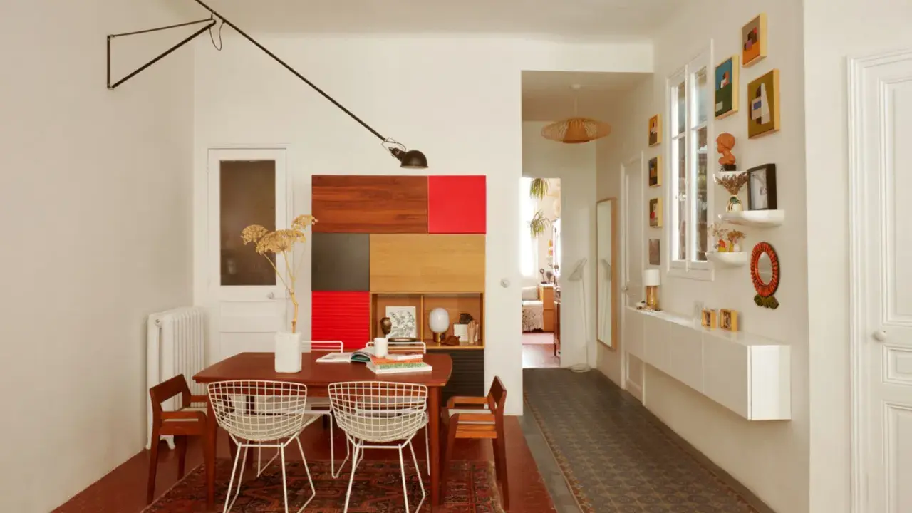 Salle à manger et couloir d'une maison ancienne, avec une table en bois, des chaises en fil de fer, une étagère colorée et des tableaux.