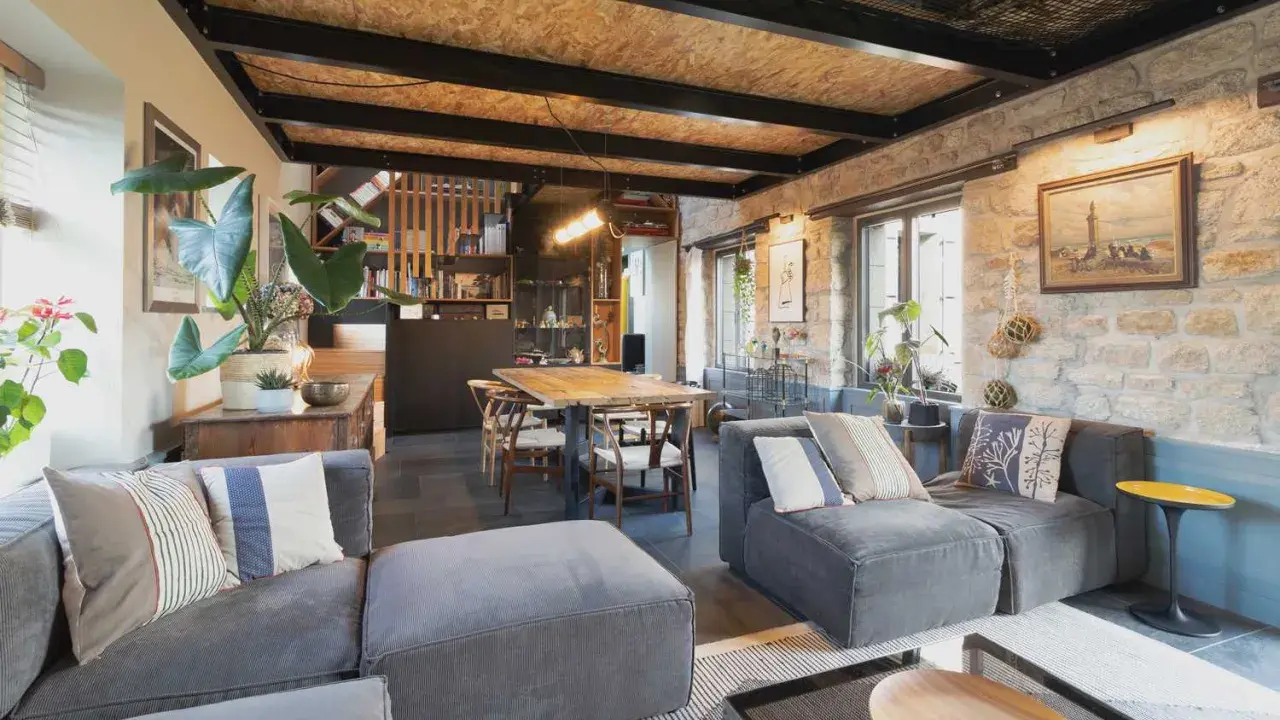 Salon cosy d'une maison de pêcheur rénovée, avec canapés gris, table en bois, murs en pierre et plantes vertes.