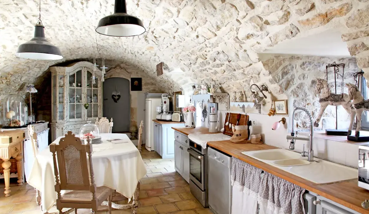 Cuisine rustique avec des vieux meubles patinés, un buffet ancien, une table dressée et des murs en pierre.