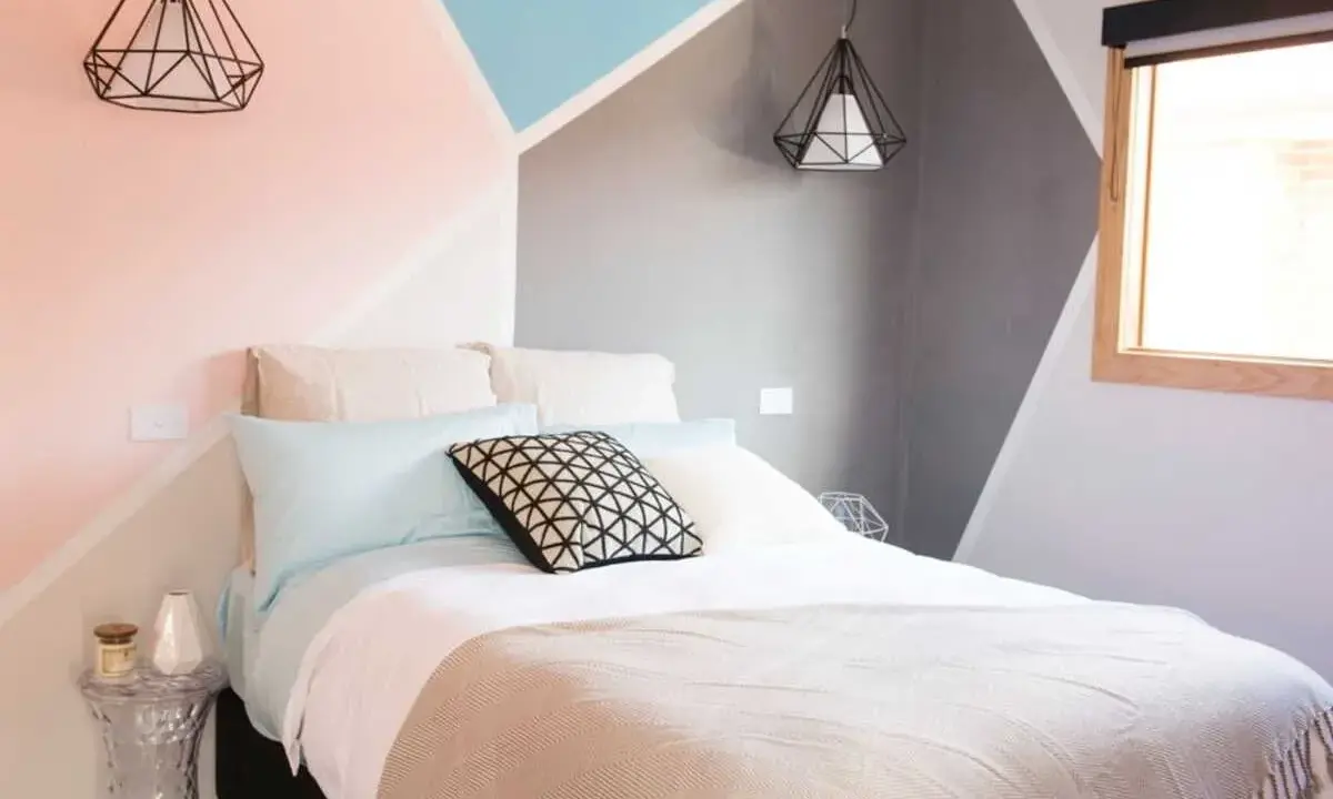 Lit moderne avec une tête de lit à peindre aux formes géométriques roses, bleues et grises. Deux suspensions noires en forme de diamant surplombent le lit.