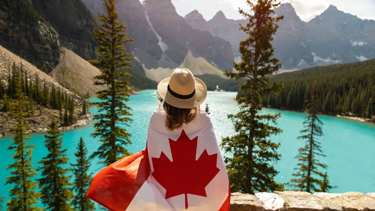 Vue d'une femme avec un drapeau canadien sur les épaules, contemplant un lac turquoise et des montagnes. Un rêve de déménagement France Canada.