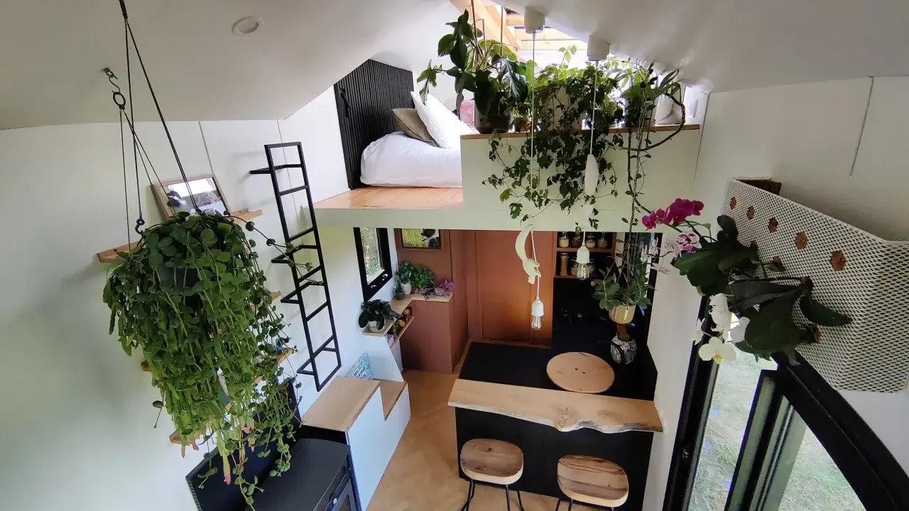 Vue aérienne du **tiny house interieur** verdoyant. Lit en mezzanine, cuisine avec bar, et cascade de plantes suspendues.