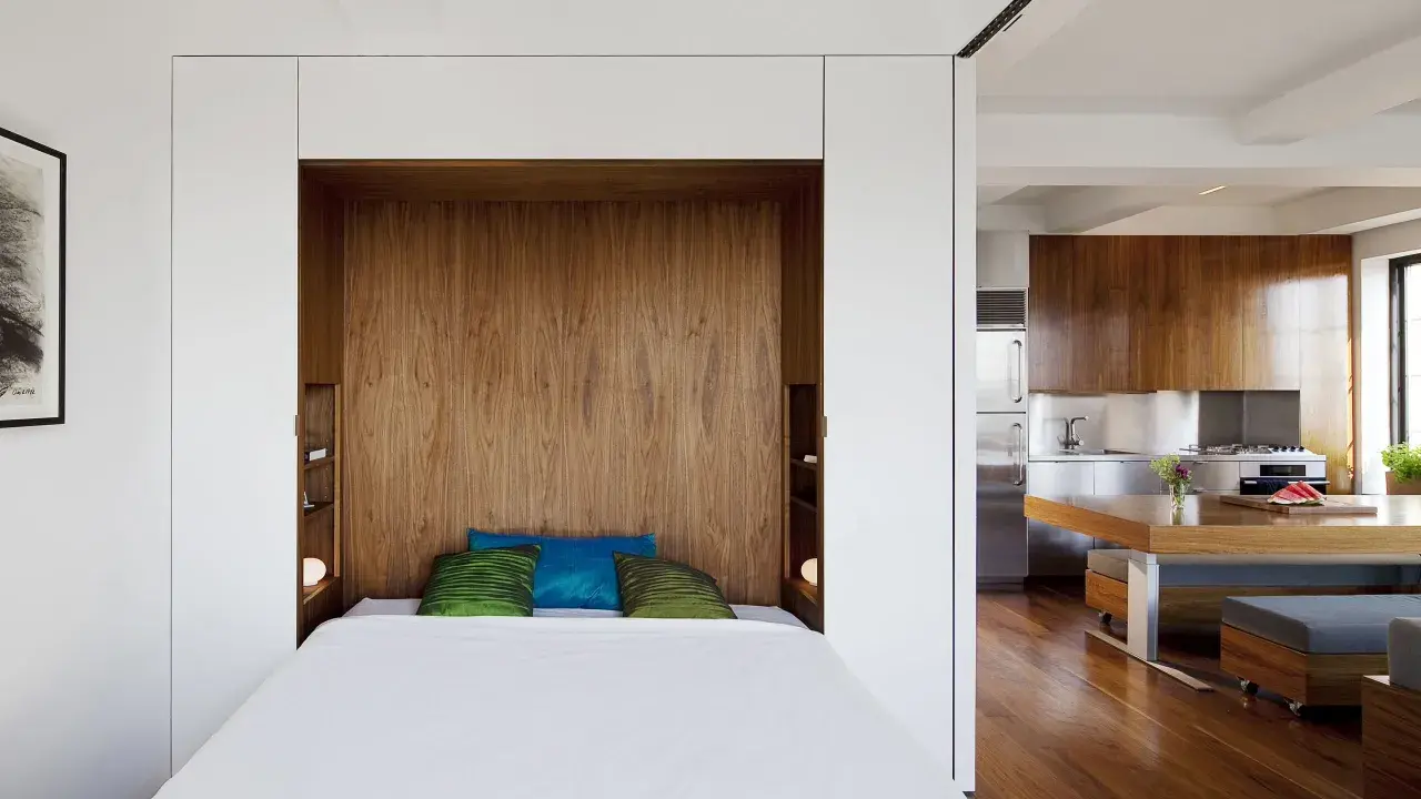Un petit espace chambre salon optimisé avec un lit escamotable en bois, des coussins colorés et une cuisine moderne adjacente.