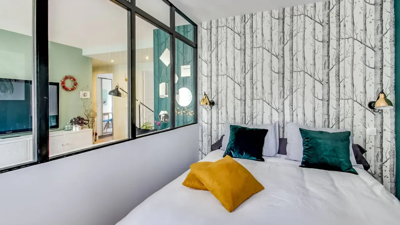 Idée pour comment amenager une chambre en longueur : un lit confortable avec des coussins colorés, un papier peint forêt et une verrière pour séparer les espaces.