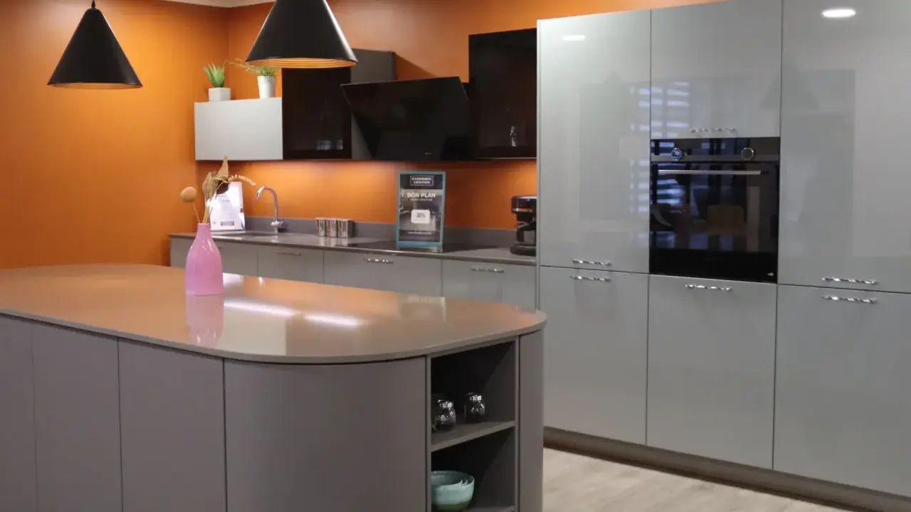 Un exemple de cuisine moderne aux lignes épurées, avec un îlot central gris et des meubles hauts gris clair. L'orange vif du mur apporte une touche de dynamisme.