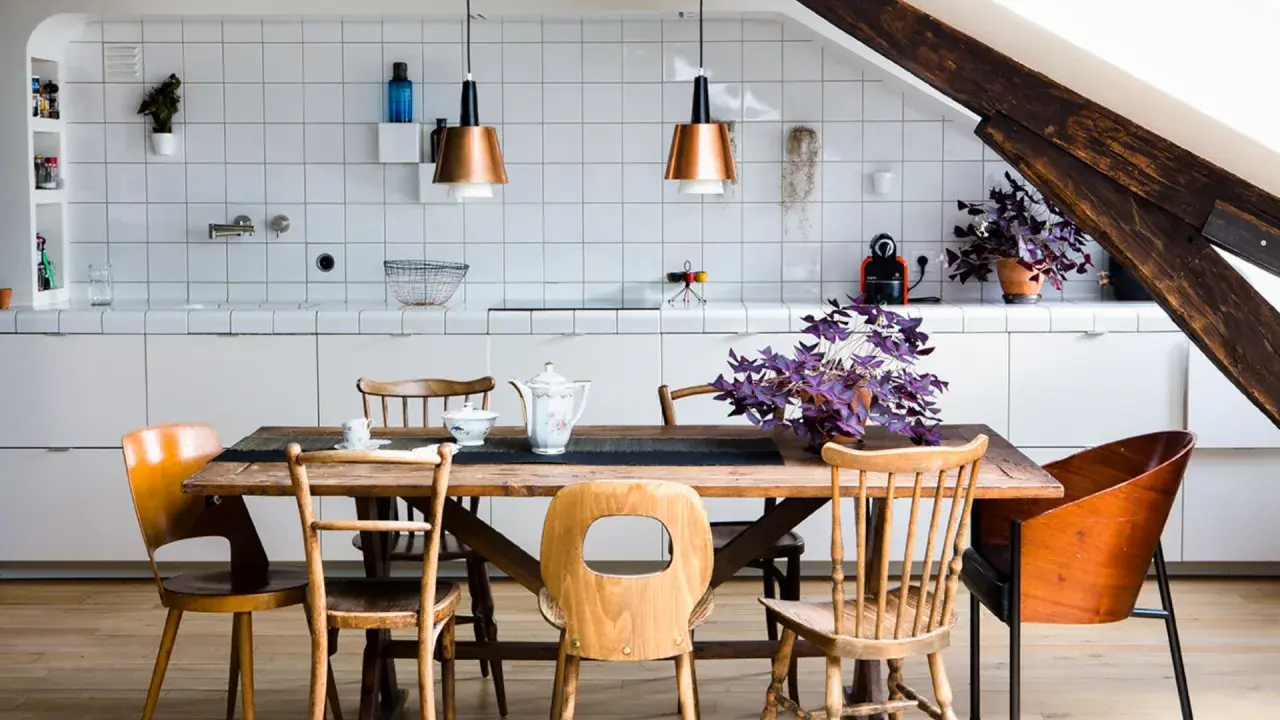 Espace de vie lumineux avec cuisine, salle à manger et salon dans la même pièce. Table en bois entourée de chaises, murs carrelés blancs, poutre de bois apparente.