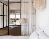 Verrière noire séparant une chambre en deux, avec une seule fenêtre visible. Un rideau blanc et un abat-jour en rotin ajoutent une touche cosy.