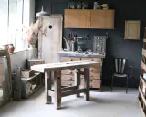 Atelier de rénovation meuble : établi en bois brut, outils, miroir ancien et étagères remplies d'objets chinés.