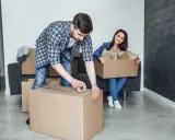 Un homme apprend comment scotcher un carton pour déménager, tandis qu'une femme assise sur un canapé tient une autre boîte remplie d'objets.