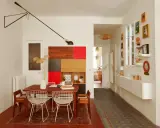 Salle à manger et couloir d'une maison ancienne, avec une table en bois, des chaises en fil de fer, une étagère colorée et des tableaux.