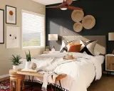 Une chambre moderne avec une décoration murale en vannerie, des tableaux abstraits et un ventilateur de plafond. Le lit est fait de blanc, avec des coussins graphiques et une touche de couleur ocre.