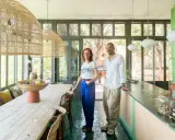 Sophie Ferjani et un homme posent dans une salle à manger lumineuse avec de grandes suspensions en rotin et un sol vert émeraude.