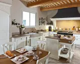 Cuisine de maison provençale avec poutres apparentes, évier en pierre, et table dressée pour un goûter gourmand.