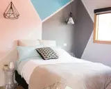 Lit moderne avec une tête de lit à peindre aux formes géométriques roses, bleues et grises. Deux suspensions noires en forme de diamant surplombent le lit.