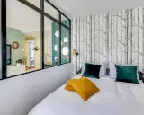 Idée pour comment amenager une chambre en longueur : un lit confortable avec des coussins colorés, un papier peint forêt et une verrière pour séparer les espaces.