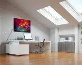 Bureau sous pente lumineux avec tableau abstrait coloré, ordinateur, plante verte et puits de lumière.