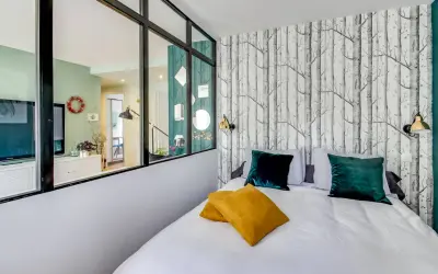 Idée pour comment amenager une chambre en longueur : un lit confortable avec des coussins colorés, un papier peint forêt et une verrière pour séparer les espaces.