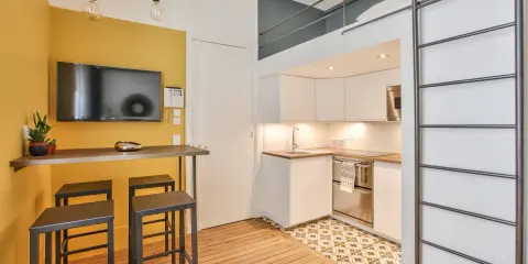 Aménagement studio 18m² : cuisine moderne, coin repas jaune vif avec TV, et lit mezzanine accessible par une échelle.