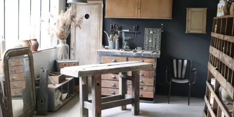 Atelier de rénovation meuble : établi en bois brut, outils, miroir ancien et étagères remplies d'objets chinés.