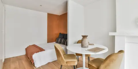Idée pour aménager studio 30m2 en 2 pièces : un coin nuit cosy avec lit et tête de lit ocre, séparé par une table ronde et des fauteuils velours.