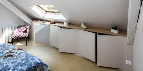 Chambre mansardée d'un coté avec lit, fauteuil à pois et rangements bas sur mesure. Un velux éclaire la pièce.