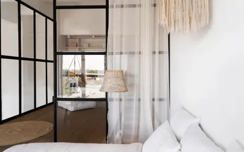 Verrière noire séparant une chambre en deux, avec une seule fenêtre visible. Un rideau blanc et un abat-jour en rotin ajoutent une touche cosy.