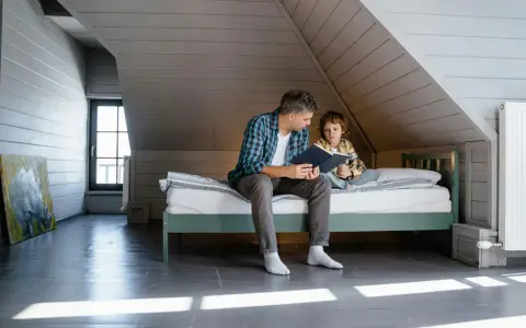 Père lisant une histoire à son fils sur un lit sous pente, dans une chambre mansardée.