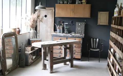 Atelier de rénovation meuble : établi en bois brut, outils, miroir ancien et étagères remplies d'objets chinés.