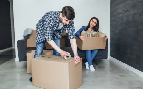 Un homme apprend comment scotcher un carton pour déménager, tandis qu'une femme assise sur un canapé tient une autre boîte remplie d'objets.