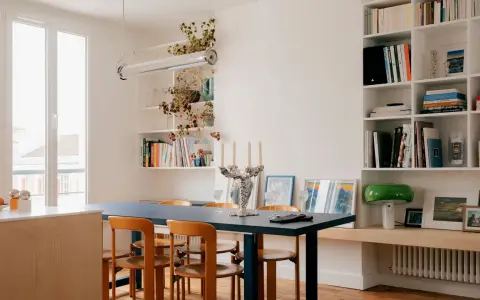 Salle à manger lumineuse d'un appartement traversant, avec table bleue, chaises en bois et étagères remplies de livres.