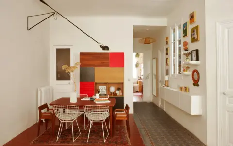 Salle à manger et couloir d'une maison ancienne, avec une table en bois, des chaises en fil de fer, une étagère colorée et des tableaux.