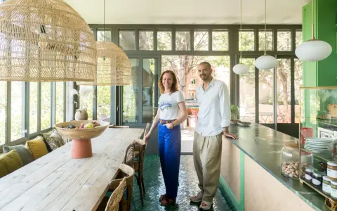 Sophie Ferjani et un homme posent dans une salle à manger lumineuse avec de grandes suspensions en rotin et un sol vert émeraude.