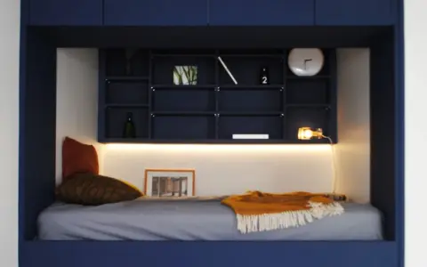 Un lit confortable dans une alcôve bleue, avec des étagères remplies d'objets décoratifs. Parfait pour refaire sa chambre avec style.