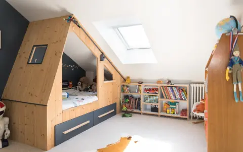 Chambre d'enfant aménagée sous les combles avec un lit cabane en bois, étagères remplies de jouets et livres. Un plan d'aménagement astucieux pour optimiser l'espace.