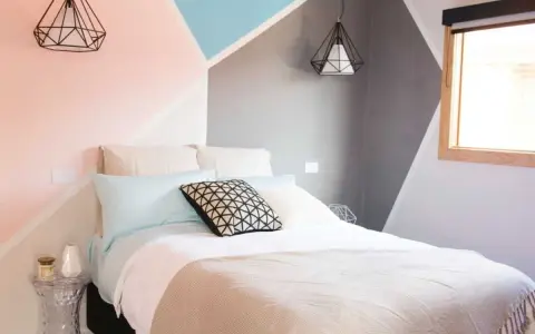Lit moderne avec une tête de lit à peindre aux formes géométriques roses, bleues et grises. Deux suspensions noires en forme de diamant surplombent le lit.