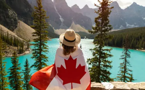 Vue d'une femme avec un drapeau canadien sur les épaules, contemplant un lac turquoise et des montagnes. Un rêve de déménagement France Canada.