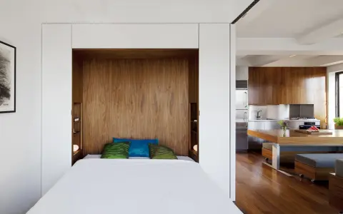 Un petit espace chambre salon optimisé avec un lit escamotable en bois, des coussins colorés et une cuisine moderne adjacente.