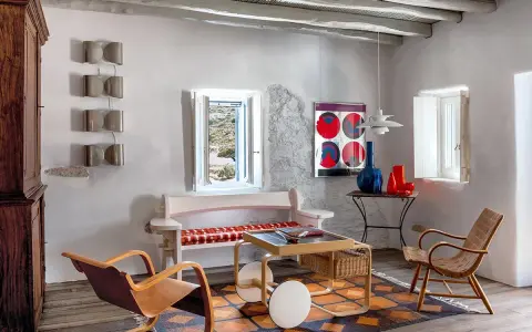 Salon d'une typique maison grecque avec mobilier en bois, tapis coloré et vue sur la mer par la fenêtre.