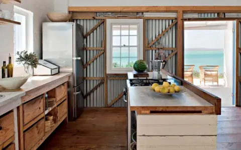 Cuisine campagne chic avec réfrigérateur, plan de travail en bois brut, murs en tôle ondulée et vue sur la mer.
