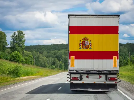 Camion aux couleurs de l'Espagne sur une route de campagne, prêt à déménager en Espagne. Paysage verdoyant sous un ciel nuageux.