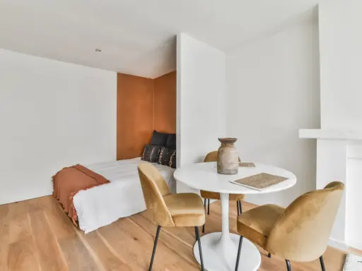 Idée pour aménager studio 30m2 en 2 pièces : un coin nuit cosy avec lit et tête de lit ocre, séparé par une table ronde et des fauteuils velours.