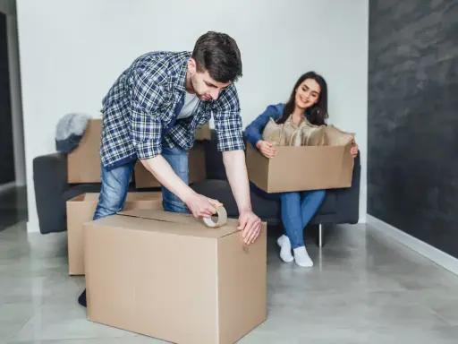 Un homme apprend comment scotcher un carton pour déménager, tandis qu'une femme assise sur un canapé tient une autre boîte remplie d'objets.