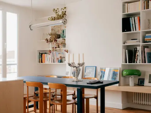 Salle à manger lumineuse d'un appartement traversant, avec table bleue, chaises en bois et étagères remplies de livres.