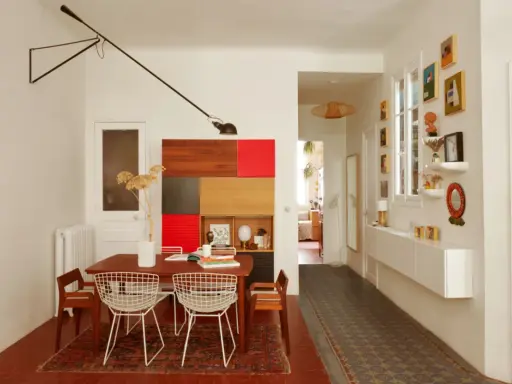 Salle à manger et couloir d'une maison ancienne, avec une table en bois, des chaises en fil de fer, une étagère colorée et des tableaux.