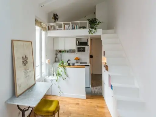 Un petit appartement optimisé avec une cuisine, un coin repas et un lit en mezzanine accessible par des escaliers blancs.