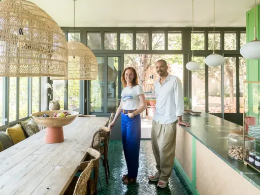 Sophie Ferjani et un homme posent dans une salle à manger lumineuse avec de grandes suspensions en rotin et un sol vert émeraude.